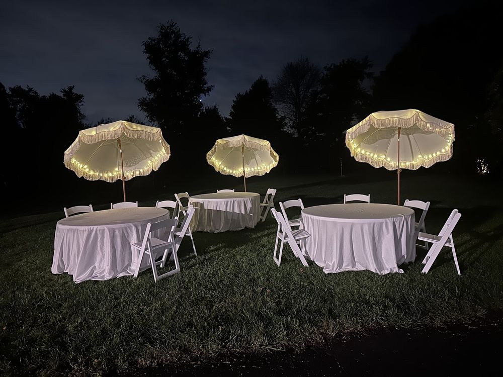 Three white umbrellas with fairy lights glowing over dressed round tables on a dark lawn